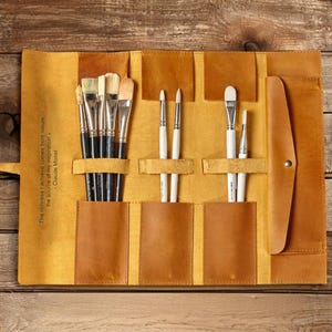 May include: A brown leather roll-up case with multiple slots for holding paint brushes. The case is open and shows a variety of brushes, including some with white bristles. The case has a quote from Claude Monet inscribed on the front: "The richness I achieve comes from nature, the source of my inspiration."