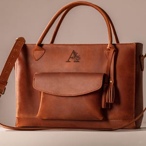 Op de afbeelding: Een bruine leren tote bag met een klepzak en een kwastje. De tas heeft twee handvatten aan de bovenkant en een afneembare schouderriem. De tas heeft een logo op de voorkant.
