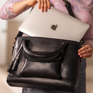 May include: Black leather satchel bag with a flap pocket and a top handle. The bag has a shoulder strap and a silver laptop inside. The laptop has the Apple logo. The initials "M|H" are embossed on the bag.