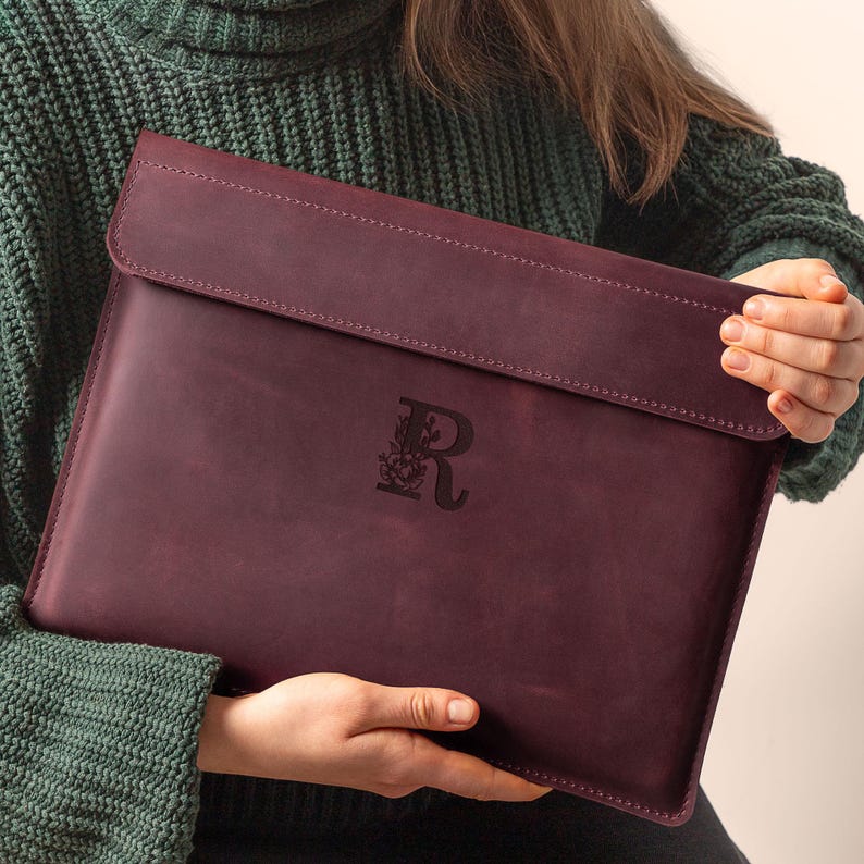 May include: A burgundy leather laptop sleeve with a flap closure. The sleeve features a debossed floral design with the letter "R" in the center. The leather has a textured appearance and is held by a person wearing a green sweater.