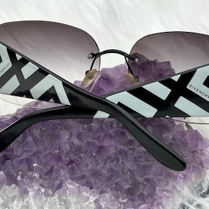 May include: Rimless sunglasses with gradient lenses. The black frames feature a geometric white and black pattern with the Givenchy logo. The temples are black. The sunglasses are resting on a purple crystal.