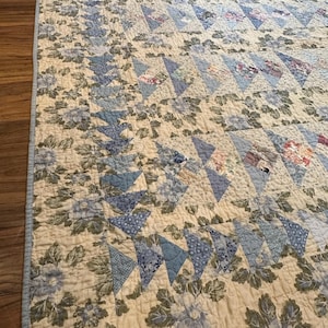 May include: A patchwork quilt featuring a cream-colored background with blue and green floral patterns. The design incorporates rows of blue fabric triangles in various shades and textures, with the text FOR EMILY stitched into the bottom corner.