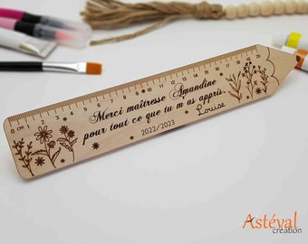 May include: A wooden ruler with engraved floral designs and text. The ruler measures up to 20 cm and includes the text "Merci maîtresse Amandine pour tout ce que tu m'as appris. Louise 2022/2023."