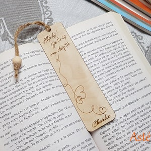 Op de afbeelding: Een houten bladwijzer met de tekst "Attends! Je finis mon chapitre" en de naam "Charlie" gegraveerd. De bladwijzer is aan een boek bevestigd met een juten touwtje. De bladwijzer heeft een hartvorm.
