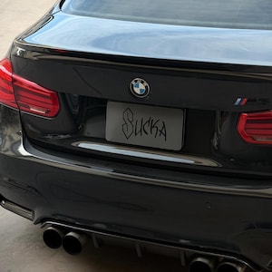 Puede incluir: Un sedán BMW M5 negro visto desde la parte trasera, con acabado brillante, luces traseras rojas, antena de aleta de tiburón y sistema de escape cuádruple. La matrícula muestra la palabra SUCKA en una fuente negra estilo grafiti sobre fondo gris.