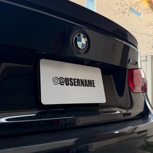 May include: Black BMW car with a white license plate. The license plate has the Instagram logo and the username "@USERNAME" in black text. The BMW logo is visible on the trunk.