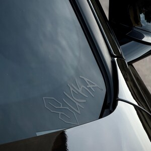 Op de afbeelding: Een close-up van de voorruit van een zwarte auto met een witte sticker met de tekst "SUCKA" in een gestileerd lettertype. De glanzende zwarte lak van de auto weerspiegelt de omgeving.