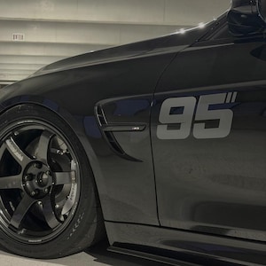 Op de afbeelding: Een close-up van de zijkant van een zwarte auto, met een grote grijze "95" sticker. De auto heeft een donkergrijze velg met een Brembo remklauw. De afbeelding benadrukt het strakke ontwerp en de sportieve uitstraling van de auto.