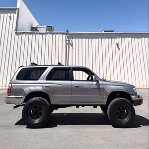 Pode incluir: Um SUV prateado com rodas pretas está estacionado em frente a um edifício branco sob um céu azul claro. O veículo tem um rack de teto e um adesivo "4 Wheel Drive".