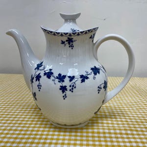 May include: White porcelain teapot with a curved spout and handle. The teapot features a blue grape vine design and a lid with a small handle. The teapot is sitting on a yellow and white checkered tablecloth.