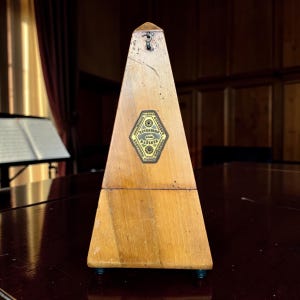May include: A pyramid-shaped wooden metronome with a light brown finish. A diamond-shaped metal plaque is centred on the front, featuring the text "METRONOME MAELZEL" and "PARIS" with decorative circular accents and a small metal hook at the top.