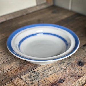 May include: A shallow ceramic bowl with a light grey interior and a wide, smooth rim. The bowl features a distinct, bold blue band encircling the outer edge of the rim, providing a clear contrast against the neutral grey base of the dish.