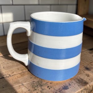 May include: A ceramic jug featuring a horizontal striped pattern in light blue and white. The vessel has a sturdy handle on the left side and a small pouring spout on the upper right rim. The interior of the jug is white and smooth.
