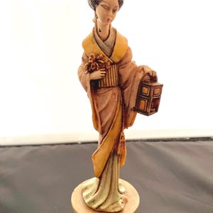 May include: A figurine of a woman in a traditional Japanese kimono holding a lantern. The figurine is made of a light brown material and is standing on a base with intricate details.