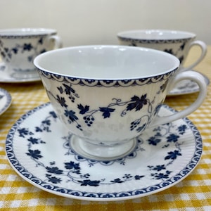 May include: A set of white teacups and saucers with a blue floral pattern. The teacups have a scalloped edge and a handle. The saucers feature a matching design. The set is displayed on a yellow and white checked tablecloth.