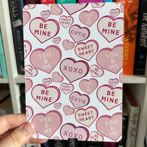 May include: A white background with pink and red heart-shaped candy conversation hearts. The hearts have various messages on them, including "Be Mine", "Sweet Heart", "You & Me", "Cutie", "XOXO", and "Love You".