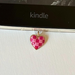 May include: A pink and red checkered heart charm with a gold-colored border hangs from a clear case. The word "kindle" is visible on the device. The background includes text from a book by Sarah Beth Durst.