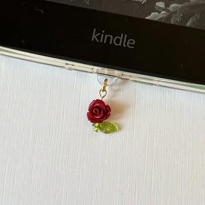 May include: A red rose charm with green leaves hangs from a gold-colored clasp. The charm is attached to a Kindle e-reader. The e-reader is black with the word "kindle" printed on it. The background is a white, textured surface.