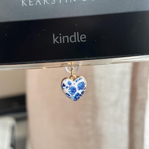 May include: A Kindle e-reader with a heart-shaped charm. The charm has a gold border and a white background with blue floral designs. The Kindle screen displays the word "kindle" and the author's name, Kearstin Dunn.