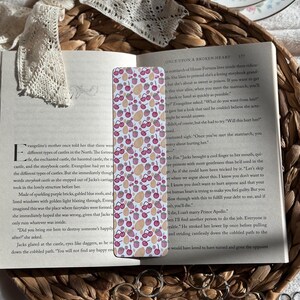Puede incluir: Un marcapáginas rectangular con un patrón de cerezas y formas de almendras en rosa, rojo y beige. El marcapáginas está colocado en un libro abierto con texto y una cinta de encaje decorativa.