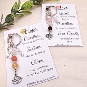 Pu&ograve; includere: Portachiavi con perline di pietre preziose e ciondoli. Un portachiavi ha un ciondolo a forma di loto e la lettera "K". L'altro portachiavi ha un ciondolo a forma di croce e cuore. Le carte elencano le pietre preziose: Moonstone, Sunstone, Citrine, Granato e Quarzo Rosa. Il testo sulle carte include "Hope" e "Love".