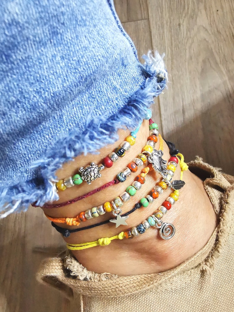 May include: Ankle bracelets with colorful beads and charms. The bracelets feature a variety of colors, including red, yellow, green, and silver. Charms include a turtle, star, feather, and spiral. The bracelets are worn with a denim jean.