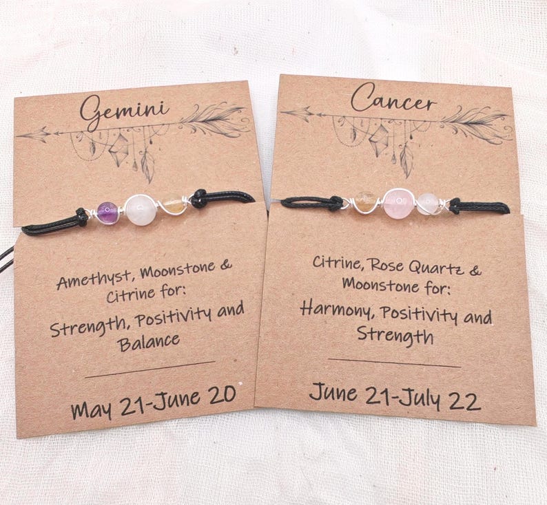 May include: Two beaded bracelets with black cords, displayed on brown cards. One card reads "Gemini" and the other "Cancer." The Gemini bracelet features amethyst, moonstone, and citrine. The Cancer bracelet features citrine, rose quartz, and moonstone.