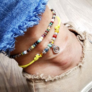 May include: Two beaded anklets with a yellow cord and a silver charm. The anklets feature colorful beads in various sizes and colors, including red, green, blue, and white. One anklet has a silver spiral charm. The anklets are worn on an ankle.