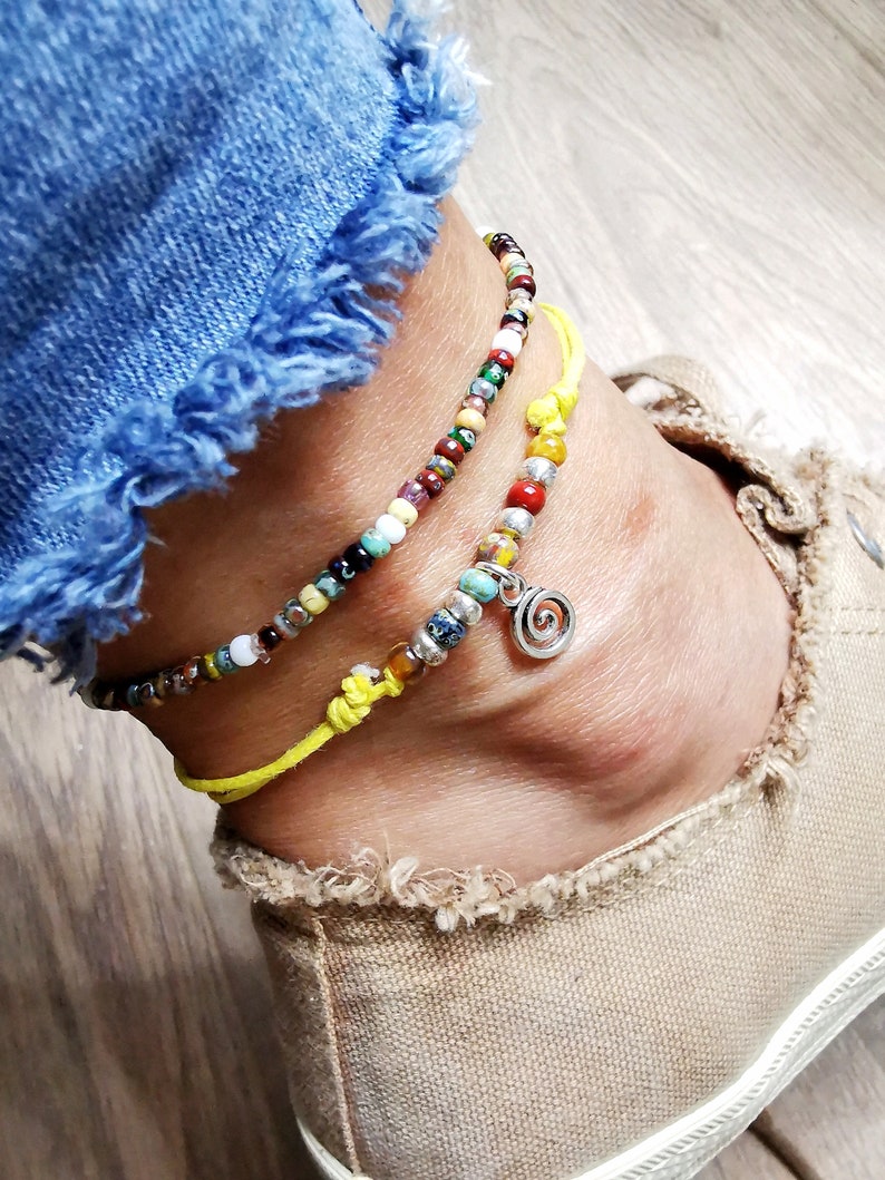 May include: Two beaded anklets with a yellow cord and a silver charm. The anklets feature colorful beads in various sizes and colors, including red, green, blue, and white. One anklet has a silver spiral charm. The anklets are worn on an ankle.