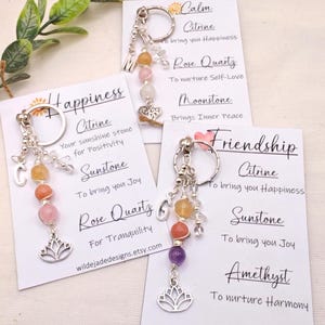 Pu&ograve; includere: Portachiavi con perline di pietre preziose arancioni, rosa e viola, attaccati a un portachiavi argentato. Ogni portachiavi ha un ciondolo e una carta con testo sulle pietre preziose. Le carte dicono "Felicit&agrave;", "Calma" e "Amicizia".
