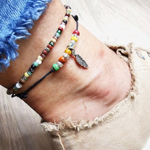 May include: Two beaded anklets. One anklet features a strand of colorful beads, while the other has a silver feather charm. The anklets are worn on an ankle, with a denim jean and canvas shoe in the background.
