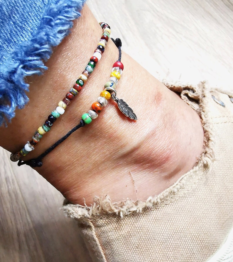 May include: Two beaded anklets. One anklet features a strand of colorful beads, while the other has a silver feather charm. The anklets are worn on an ankle, with a denim jean and canvas shoe in the background.