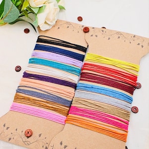 May include: Two spools of colorful elastic cord in various colors, including black, blue, purple, white, pink, orange, and yellow. The cords are neatly wound on decorative cardboard spools with wooden button accents. A floral arrangement is in the background.
