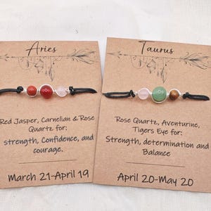 May include: Two beaded bracelets on brown cards, one for Aries and one for Taurus. The Aries bracelet has red and pink beads. The Taurus bracelet has pink, green, and brown beads. Text on the cards describes the zodiac signs.