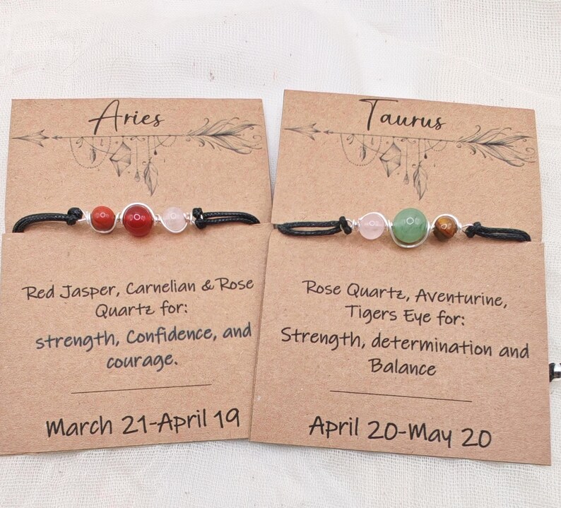 May include: Two beaded bracelets on brown cards, one for Aries and one for Taurus. The Aries bracelet has red and pink beads. The Taurus bracelet has pink, green, and brown beads. Text on the cards describes the zodiac signs.