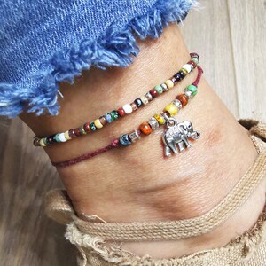 May include: Two beaded anklets. The top anklet features colorful beads in various colors, while the bottom one is a deep red with a silver elephant charm. The anklets are worn on an ankle with a denim jean and tan shoe in the background.