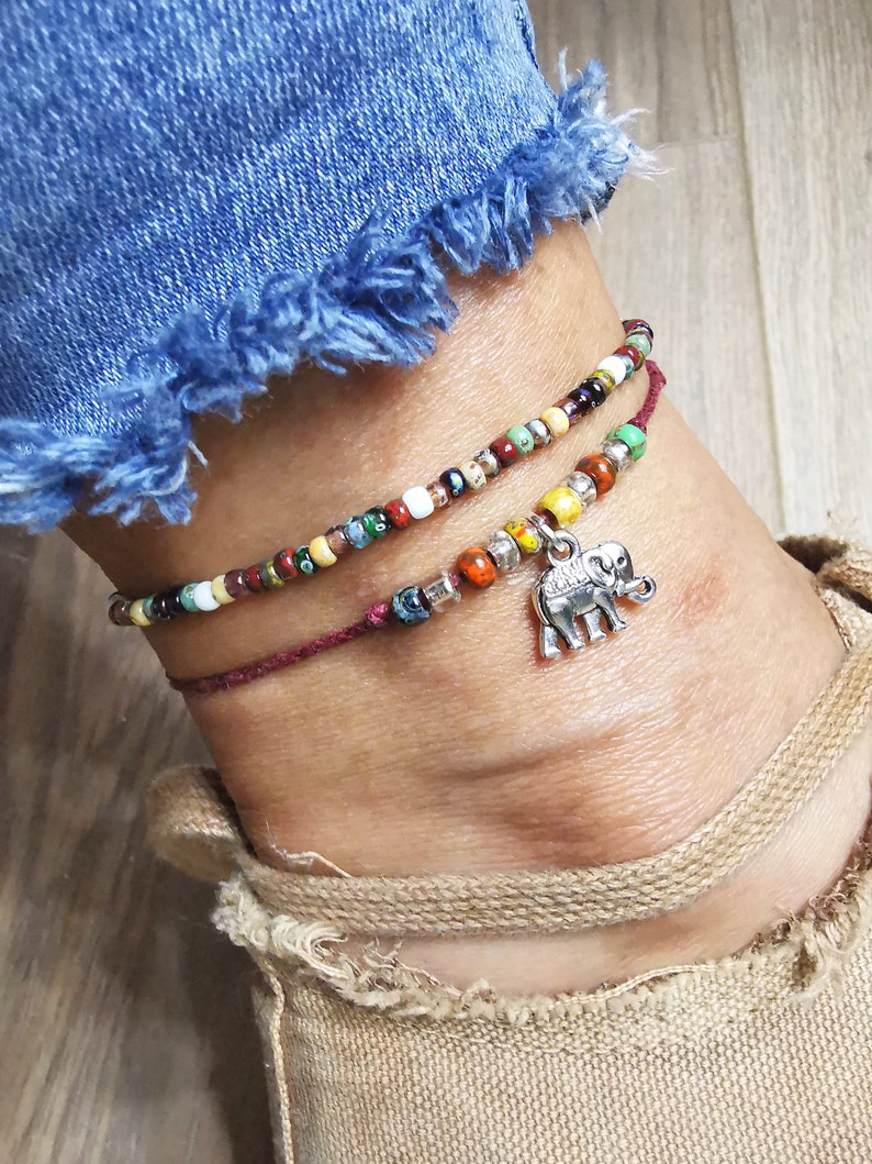 May include: Two beaded anklets. The top anklet features colorful beads in various colors, while the bottom one is a deep red with a silver elephant charm. The anklets are worn on an ankle with a denim jean and tan shoe in the background.