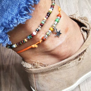 May include: Two beaded anklets. One is multi-colored with a variety of beads. The other is orange with a silver star charm. The anklets are worn on an ankle with a canvas shoe and blue jeans.