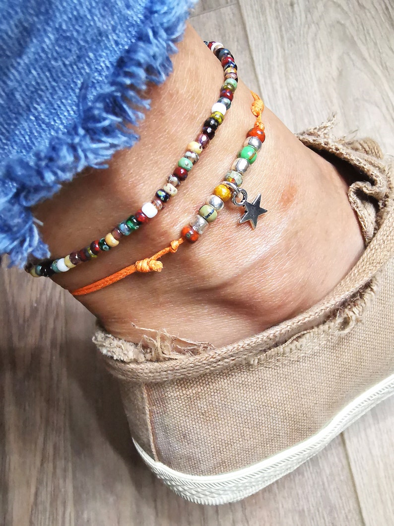 May include: Two beaded anklets. One is multi-colored with a variety of beads. The other is orange with a silver star charm. The anklets are worn on an ankle with a canvas shoe and blue jeans.