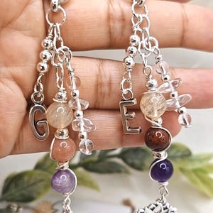Pu&ograve; includere: Orecchini in argento con perline e ciondoli di pietre preziose. Ogni orecchino presenta una catena con un ciondolo a lettera, perline di pietre preziose in vari colori e un ciondolo a forma di fiore di loto. Gli orecchini hanno un tema spirituale.