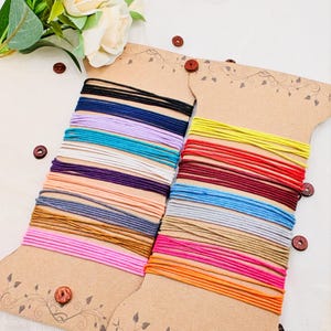 May include: Two spools of colorful elastic cord in various colors, including black, blue, purple, and pink. The cords are neatly wound on decorative cardboard spools with wooden button accents. A floral arrangement is in the background.