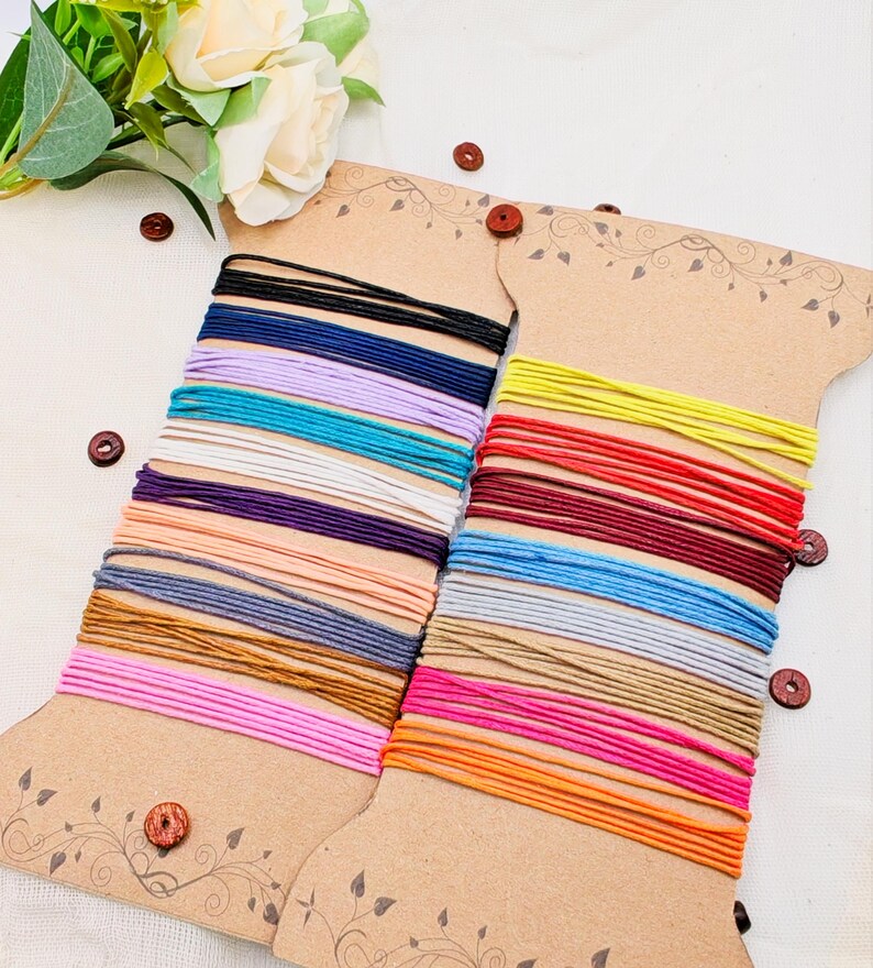 May include: Two spools of colorful elastic cord in various colors, including black, blue, purple, and pink. The cords are neatly wound on decorative cardboard spools with wooden button accents. A floral arrangement is in the background.