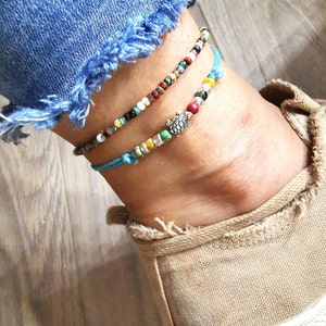 May include: Two beaded anklets. One anklet features a small silver turtle charm. The anklets are made with colorful beads and blue string. The anklets are worn on an ankle with a pair of blue jeans and a tan shoe.