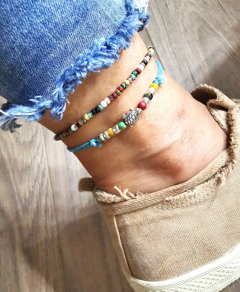 May include: Two beaded anklets. One anklet features a small silver turtle charm. The anklets are made with colorful beads and blue string. The anklets are worn on an ankle with a pair of blue jeans and a tan shoe.