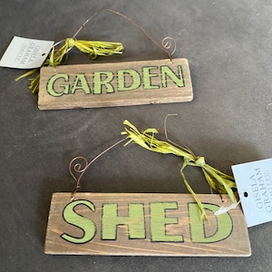May include: Two wooden signs with green lettering, one says "GARDEN" and the other says "SHED". Both signs have a wire hanger and a green raffia bow.
