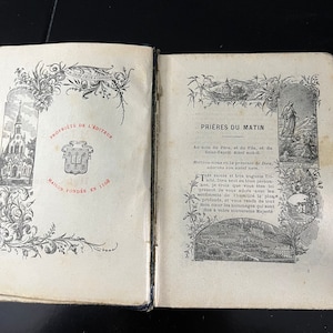 May include: An open book with a black cover and gold accents. The book is open to a page with a black and white illustration of a church and the text "PRIÈRES DU MATIN". The text "PROPRIETE DE L'EDITEUR" and "MAISON FONDÉE EN 1468" are visible on the left page.