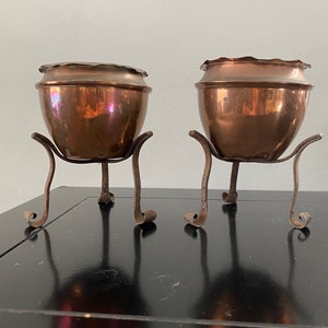May include: Two copper plant pots with a hammered finish, each sitting on a black metal stand with three legs.