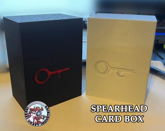 May include: Two 3D printed card boxes, one black and one white, with a stylized key design on the front. The black box has a red key design and the white box has a white key design. The white box has the text "SPEARHEAD CARD BOX" printed on the front.