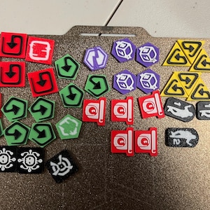 Pode incluir: Uma coleção de fichas de jogo coloridas em várias formas, incluindo quadrados, pentágonos e triângulos. As fichas apresentam símbolos como setas, dados e criaturas estilizadas. A paleta de cores inclui vermelho, verde, roxo, amarelo e preto.