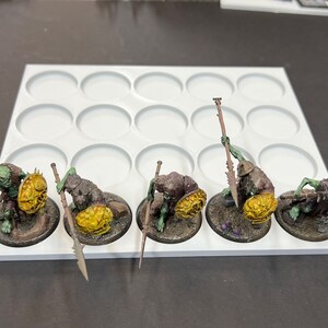 Puede incluir: Cinco figuras en miniatura de color verde y marr&oacute;n con escudos amarillos y lanzas se muestran en una base de pl&aacute;stico blanca con 12 c&iacute;rculos redondos.